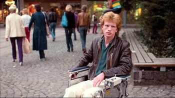 Movie still from “Spetters” (1980), directed by Paul Verhoeven – A man sitting in a wheelchair on the sidewalk; Medium shot, Low angle
