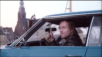 Movie still from “Spetters” (1980), directed by Paul Verhoeven – A man sitting in the driver's seat of a blue car; Medium shot, Over the shoulder angle