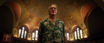 Movie still from “Spiral” (2021), directed by Darren Lynn Bousman – A man wearing glasses standing in front of stained glass windows; Medium shot, Low angle