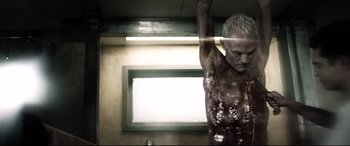 Movie still from “Spiral” (2021), directed by Darren Lynn Bousman – A shirtless man with no shirt hanging in a room; Medium shot, Low angle