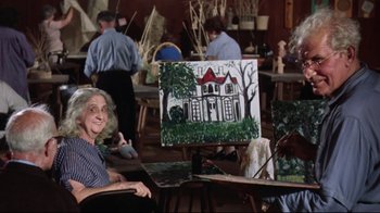 An older woman sitting in front of an easel painting a house
