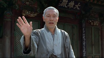 An older man wearing a robe is holding up three fingers