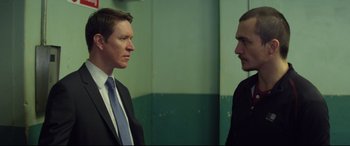 Movie still from “Starred Up” (2013), directed by David Mackenzie – Two men in business suits standing next to each other; Medium shot, Over the shoulder angle