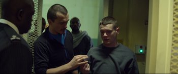 Movie still from “Starred Up” (2013), directed by David Mackenzie – A man is getting his ear pierced by another man; Medium shot, Over the shoulder angle