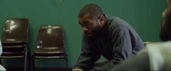 Movie still from “Starred Up” (2013), directed by David Mackenzie – A man leaning on a chair in front of a green wall; Medium shot, Over the shoulder angle
