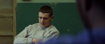 Movie still from “Starred Up” (2013), directed by David Mackenzie – A man sitting in front of a green wall with his arms crossed; Medium shot, Over the shoulder angle