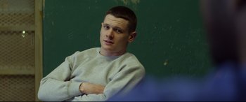 Movie still from “Starred Up” (2013), directed by David Mackenzie – A man sitting in front of a green wall with his arms crossed; Medium shot, Over the shoulder angle
