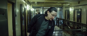 Movie still from “Starred Up” (2013), directed by David Mackenzie – A man with long black hair wearing glasses and a black jacket; Medium shot, Over the shoulder angle