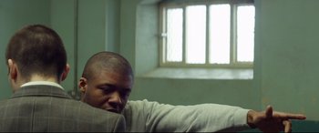 Movie still from “Starred Up” (2013), directed by David Mackenzie – A man sitting on a couch in front of an open window; Medium shot, Over the shoulder angle