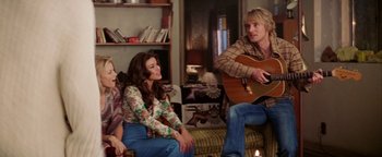Movie still from “Starsky & Hutch” (2004), directed by Todd Phillips – A group of people sitting around a living room playing a guitar; Medium shot, Over the shoulder angle