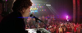 Movie still from “Starsky & Hutch” (2004), directed by Todd Phillips – A man is speaking at a microphone in front of a crowd of onlookers; Medium shot, Low angle