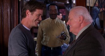 Movie still from “State and Main” (2000), directed by David Mamet – Two men are talking to each other in front of another man holding a cell phone; Medium shot, Over the shoulder angle