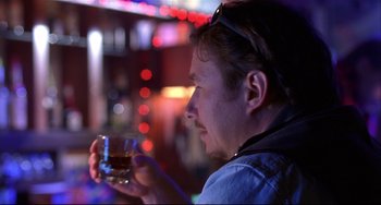 Movie still from “State and Main” (2000), directed by David Mamet – A man holding a glass of alcohol in his hand; Close Up shot, Over the shoulder angle
