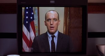 Movie still from “State and Main” (2000), directed by David Mamet – A man wearing a suit and tie in front of an american flag on a television screen; Close Up shot, Low angle