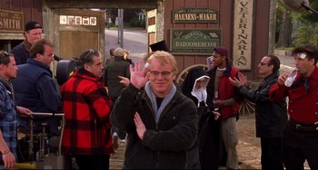Movie still from “State and Main” (2000), directed by David Mamet – A man waving to the camera while standing in front of a building; Medium shot, Over the shoulder angle