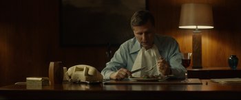 Movie still from “Stockholm” (2018), directed by Robert Budreau – A man sitting at a table with a plate of food; Medium shot, Low angle