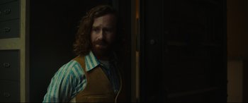 Movie still from “Stockholm” (2018), directed by Robert Budreau – A man with long hair and a beard standing next to a door; Medium shot, Over the shoulder angle