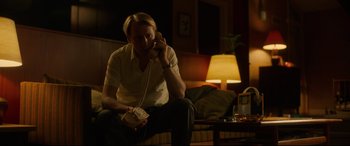 Movie still from “Stockholm” (2018), directed by Robert Budreau – A man sitting on a couch talking on a phone; Medium shot, Low angle