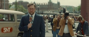 Movie still from “Stockholm” (2018), directed by Robert Budreau – A man in a suit and tie holding a microphone; Medium shot, Over the shoulder angle