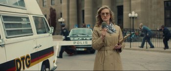 Movie still from “Stockholm” (2018), directed by Robert Budreau – A woman is standing in front of a police car and holding a microphone; Medium shot, Low angle