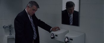 Movie still from “Stone” (2010), directed by John Curran – A man in a suit and tie touching a urinal in a restroom; Medium shot, High angle