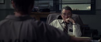 Movie still from “Stone” (2010), directed by John Curran – An older man sitting in an office chair with his hand under his chin; Medium shot, Over the shoulder angle