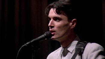 Movie still from “Stop Making Sense” (1984), directed by Jonathan Demme – A man is singing into a microphone in front of a crowd; Close Up shot, Low angle