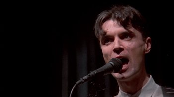 Movie still from “Stop Making Sense” (1984), directed by Jonathan Demme – A man is singing into a microphone on a stage; Close Up shot, Low angle