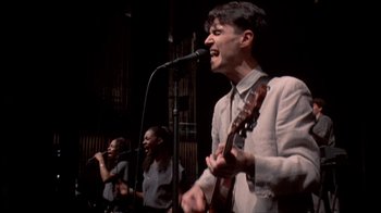 Movie still from “Stop Making Sense” (1984), directed by Jonathan Demme – A group of people that are singing on stage; Medium shot, Low angle