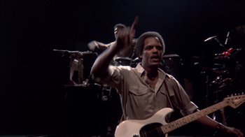 Movie still from “Stop Making Sense” (1984), directed by Jonathan Demme – A man holding a guitar in front of a band; Medium shot, Low angle