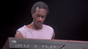 Movie still from “Stop Making Sense” (1984), directed by Jonathan Demme – A man sitting at a keyboard in front of a microphone; Close Up shot, Low angle