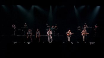 Movie still from “Stop Making Sense” (1984), directed by Jonathan Demme – A group of people on a stage singing and playing a guitar; Extreme Wide shot, High angle
