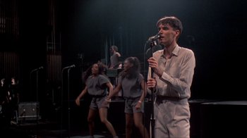 Movie still from “Stop Making Sense” (1984), directed by Jonathan Demme – A group of people on a stage with a microphone; Medium shot, High angle
