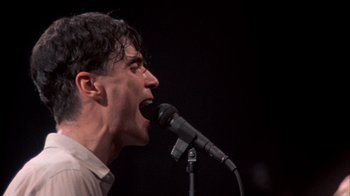 Movie still from “Stop Making Sense” (1984), directed by Jonathan Demme – A man is singing into a microphone on a stage; Close Up shot, Low angle
