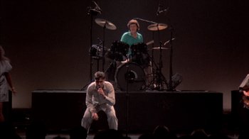 Movie still from “Stop Making Sense” (1984), directed by Jonathan Demme – A man sitting on top of a stage next to a man playing drums; Wide shot, High angle