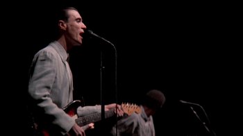 Movie still from “Stop Making Sense” (1984), directed by Jonathan Demme – A man singing into a microphone on a stage; Medium shot, Low angle