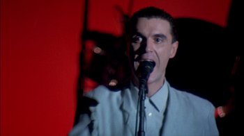 Movie still from “Stop Making Sense” (1984), directed by Jonathan Demme – A man is speaking into a microphone in front of a red wall; Close Up shot, Low angle