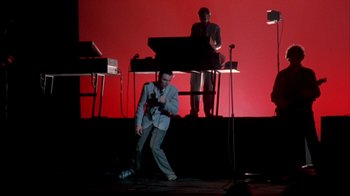 Movie still from “Stop Making Sense” (1984), directed by Jonathan Demme – A man sitting on a stage in front of a keyboard; Wide shot, Low angle