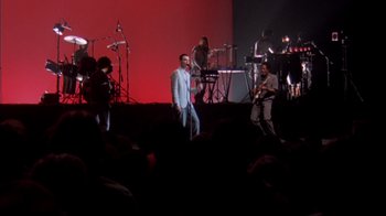 Movie still from “Stop Making Sense” (1984), directed by Jonathan Demme – A group of people on a stage with a microphone; Extreme Wide shot, High angle