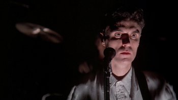 Movie still from “Stop Making Sense” (1984), directed by Jonathan Demme – A man standing in front of a microphone in a dark room; Close Up shot, Low angle