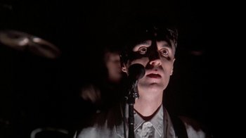 Movie still from “Stop Making Sense” (1984), directed by Jonathan Demme – A man standing in front of a microphone in a dark room; Close Up shot, Low angle