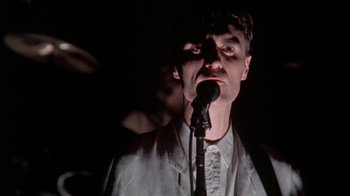 Movie still from “Stop Making Sense” (1984), directed by Jonathan Demme – A man in a white suit is singing into a microphone; Close Up shot, Low angle