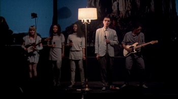 Movie still from “Stop Making Sense” (1984), directed by Jonathan Demme – A group of people standing on a stage with microphones; Wide shot, Low angle