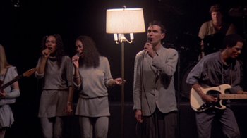 Movie still from “Stop Making Sense” (1984), directed by Jonathan Demme – A group of people standing in front of a microphone; Medium shot, High angle