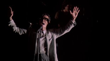 Movie still from “Stop Making Sense” (1984), directed by Jonathan Demme – A man in a suit and glasses singing into a microphone; Medium shot, Low angle