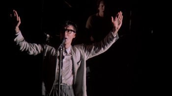 Movie still from “Stop Making Sense” (1984), directed by Jonathan Demme – A man with a microphone in front of a microphone stand; Medium shot, Low angle