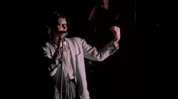 Movie still from “Stop Making Sense” (1984), directed by Jonathan Demme – A man in a white jacket is singing into a microphone; Medium shot, Low angle