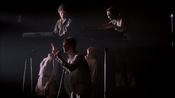 Movie still from “Stop Making Sense” (1984), directed by Jonathan Demme – A group of people standing around a stage; Wide shot, Low angle