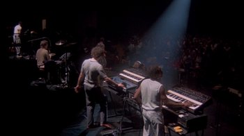 Movie still from “Stop Making Sense” (1984), directed by Jonathan Demme – A group of men playing keyboards on a stage; Wide shot, High angle