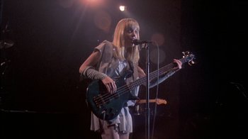 Movie still from “Stop Making Sense” (1984), directed by Jonathan Demme – A woman holding a guitar and singing into a microphone; Medium shot, Low angle